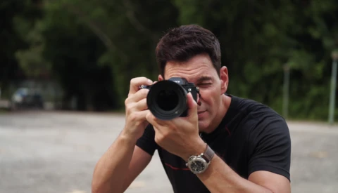 Male photographer holding a DSLR camera with telephoto lens, looking through the viewfinder.