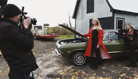 Photographer shooting a woman in a red coat posing by a vintage car with open hood.