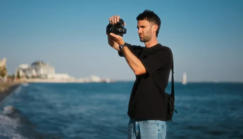 Man in black shirt holding a compact camera up to his face while standing on a rocky beach with clear blue ocean and coastal buildings in the background.