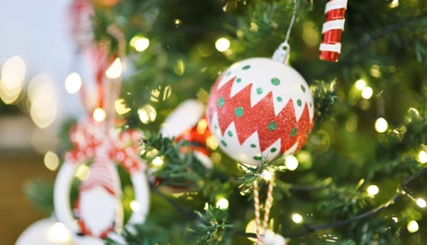 Red and white patterned ornament hanging from evergreen branch with warm bokeh lights in background.