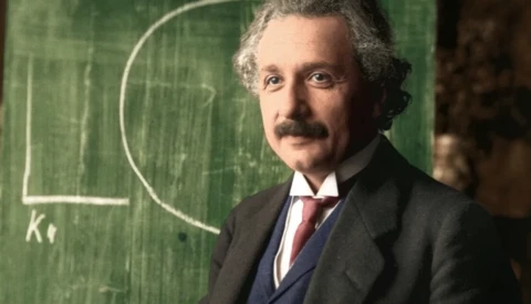 Man in formal attire with mustache standing before green chalkboard with geometric diagram.