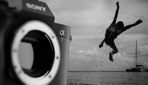 Sony α7 camera body in foreground with silhouetted person diving into water near sailboat in background, black and white.