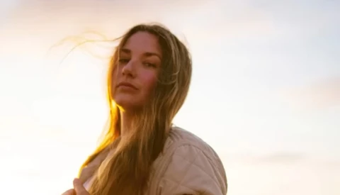 Woman with long blonde hair backlit by warm golden sunset light against a soft, overcast sky.