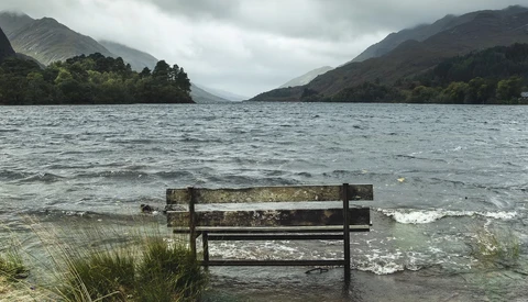 Weathered wooden bench partially submerged in calm lake water, facing mountains under overcast sky.