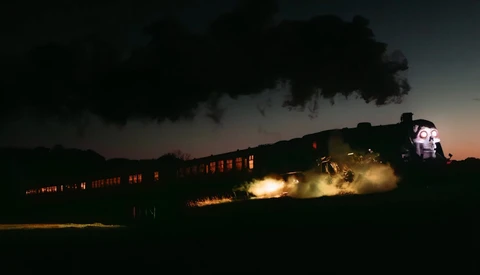 Steam locomotive at night with illuminated passenger cars and dramatic smoke plume against darkening sky.
