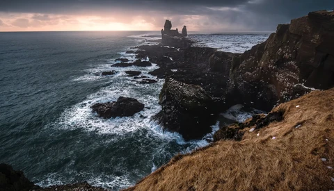 Dramatic coastal landscape with jagged rock formations and ruins silhouetted against a moody dawn sky.