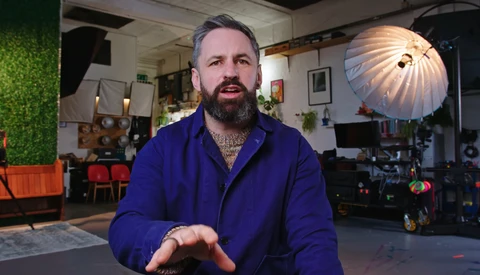 Man with beard wearing blue jacket speaks directly to camera in professional studio setting.