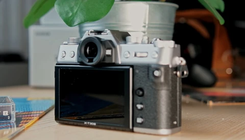 Rangefinder camera with articulating rear screen displayed on wooden desk with potted plant in background.