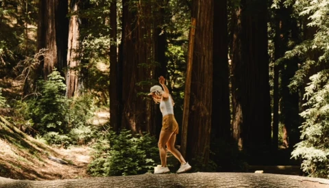 Woman in tan pants and white top jumping on a forest trail surrounded by towering redwood trees.