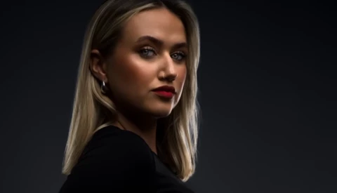 Woman with shoulder-length blonde hair wearing dark clothing, photographed against a dark background in profile.