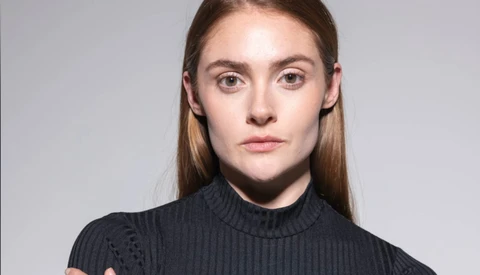 Portrait of a young woman with long brown hair wearing a dark turtleneck against a neutral gray background.