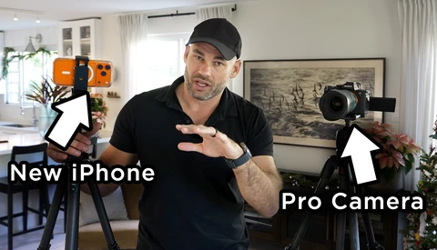 Content creator in black cap and shirt gestures between a smartphone on a tripod and a professional cinema camera, both labeled with white arrows in a bright home studio.
