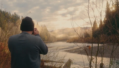 Photographer shooting a misty river landscape at golden hour with forested banks and bare branches in foreground.