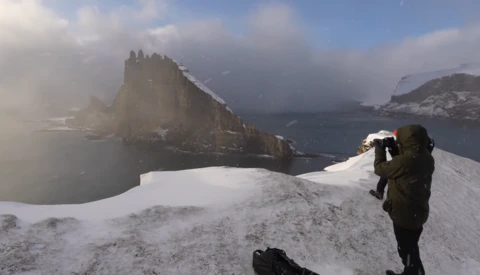 Photographer with telephoto lens on snowy ridge overlooking dramatic rocky peak and misty fjord landscape.