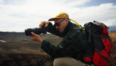 Photographer in yellow hard hat and backpack crouches while composing a shot with a telephoto lens in an arid landscape.