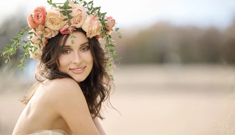 Woman wearing a floral crown with peach and coral roses, photographed outdoors with soft natural backlighting.