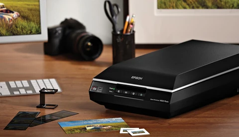 Flatbed scanner on wooden desk with photo prints, camera, and studio setup visible in background.