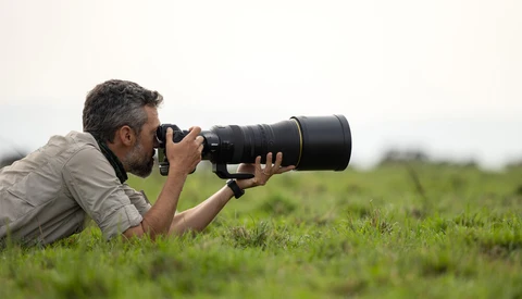 Wildlife photographer lying prone in grass, holding telephoto lens while composing a shot across an open field.