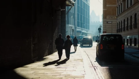 Sunlit urban street with pedestrians and parked vehicles casting long shadows on pavement.