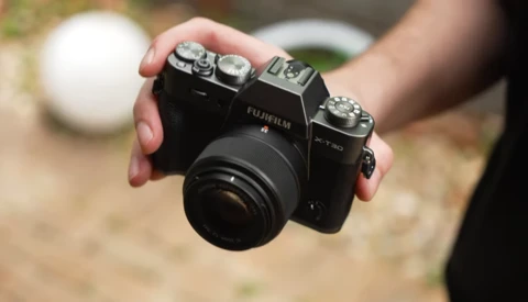 Hand holding a Fujifilm X-T30 mirrorless camera with attached lens outdoors.