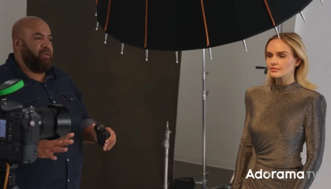 Cinematographer operating camera while interviewing woman under large softbox lighting setup.