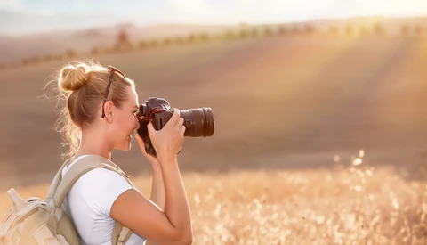 Female photographer holding a DSLR camera up to her face while standing in a golden wheat field at sunset.