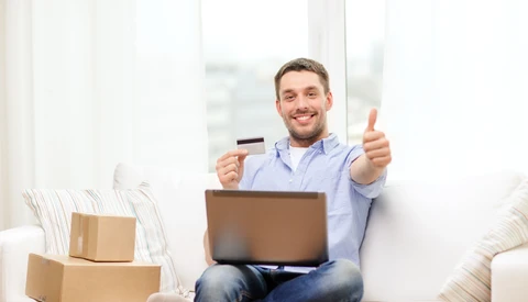 Man sitting on a couch holding a credit card and laptop while giving a thumbs up.