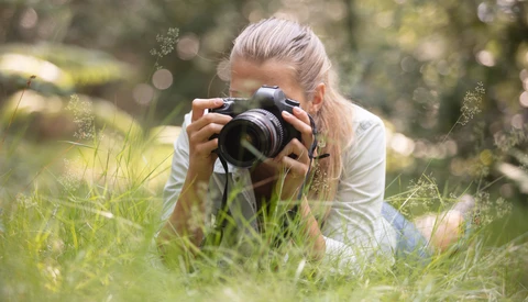 Photographer crouching in tall grass, holding a DSLR camera up to her face while composing a shot.