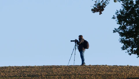 Photographer standing on a ridge silhouetted against blue sky, looking through a telephoto lens mounted on a tripod.