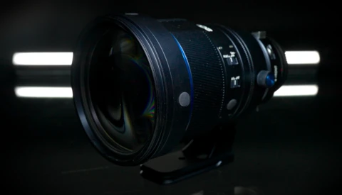 Wide-angle camera lens with blue focus ring, backlit by studio lights against dark background.