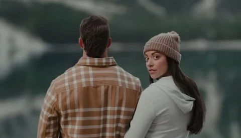 Two people standing outdoors by a lake, one wearing a tan plaid shirt facing away and one wearing a cream hoodie and beanie looking toward camera.