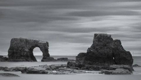 Rock formations with natural arch rising from shallow ocean water under dramatic cloudy sky.