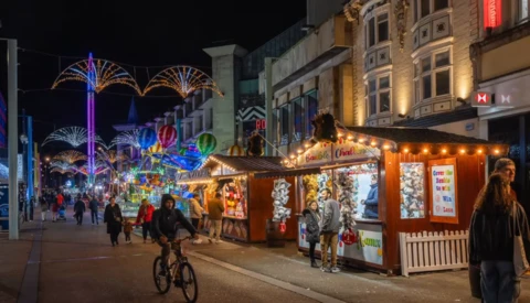 Nighttime holiday market street with illuminated vendor stalls, decorative light installations, and pedestrians shopping.