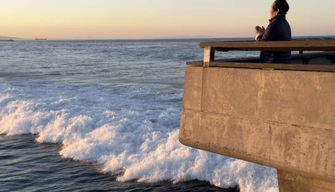 Person sitting on concrete pier overlooking ocean waves at dusk.