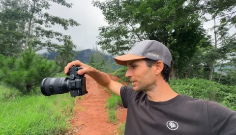 Photographer in gray cap holds mirrorless camera up to eye while standing on red dirt path surrounded by lush green vegetation and distant mountains.