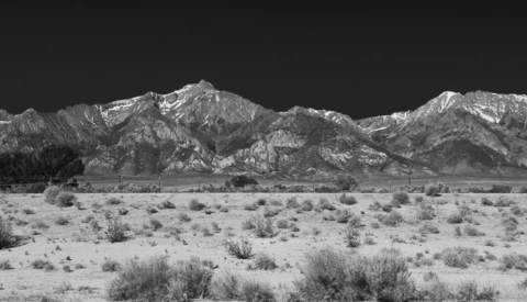 Snow-capped mountain range rising above sparse desert scrubland under dark sky.