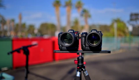 Two cinema cameras mounted on a rig against a blurred outdoor background with palm trees and red equipment.