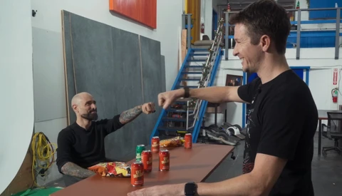 Two men in a workshop demonstrate a lighting or photography technique using colorful cans and a chalkboard backdrop.