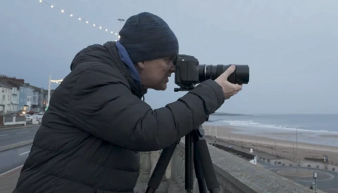 Photographer using a telephoto lens on a tripod while positioned on an elevated walkway overlooking a beach.