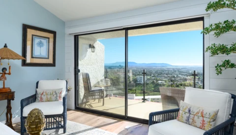 Bright bedroom with open sliding glass doors revealing a sprawling mountain valley landscape and clear blue sky.