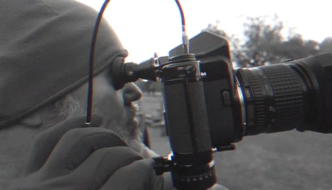 Photographer holding a camera with telephoto lens up to their eye, viewed from the side profile outdoors.