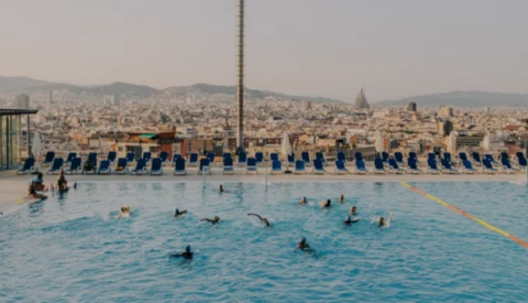 Swimmers in a bright blue public pool with a dense cityscape and mountains visible in the background.