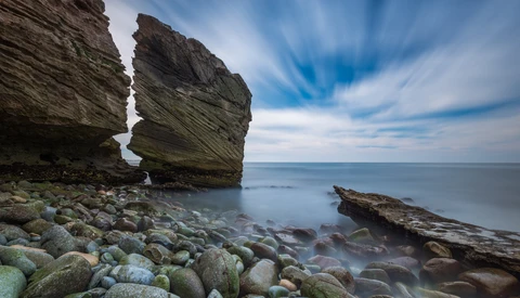 Dramatic coastal rock formations with layered striations separated by a narrow gap, surrounded by boulders and still water under blue sky.
