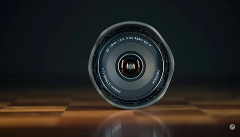 Camera lens viewed head-on against a dark background, positioned on a reflective wooden surface.