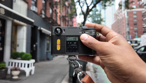 Hand holding a rangefinder film camera with yellow accent on the body, mounted on a tripod in an urban street setting.