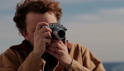 Person holding a rangefinder film camera up to their face, photographing against a coastal backdrop.