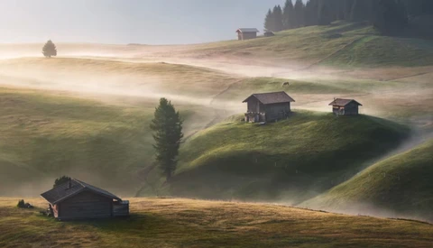 Alpine valley at sunrise with scattered wooden chalets nestled on misty hillsides surrounded by evergreen forests.