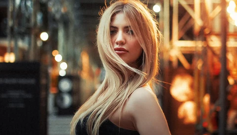 Woman with long blonde hair looking over shoulder in urban evening setting with warm golden lighting.