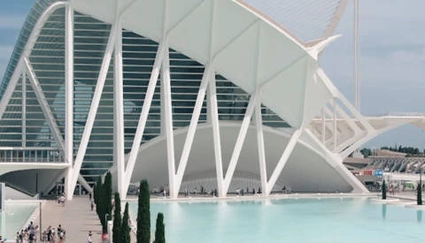 Modern white architectural structure with curved ribbed exterior beside turquoise reflecting pool, cypress trees, and glass building facade.