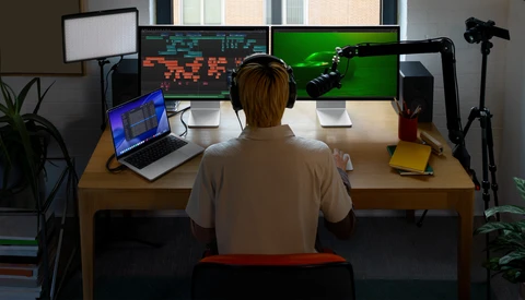Person seated at wooden desk viewing multiple monitors displaying game graphics and code, with laptop, microphone, and lighting equipment visible.